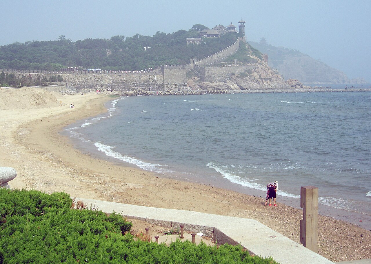 A wide curve of pale sand beach with surf, a person standing near the water, and a long defensive wall climbing the headland beyond