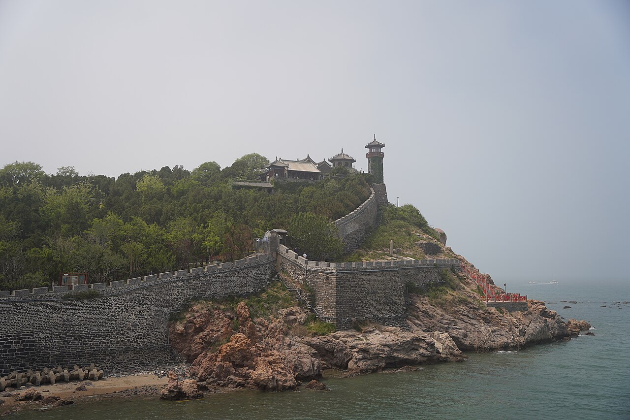 The headland seen across the water from the south, with a crenellated wall at the shore, rocks at its base and trees and halls above in hazy air