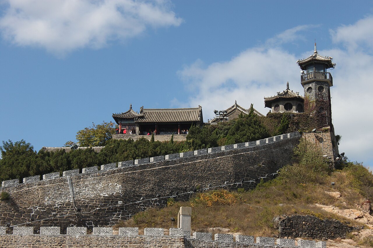 A cluster of dark-roofed temple halls and a stone watchtower on a headland above a crenellated wall, with a green slope in front and blue sky with cloud behind
