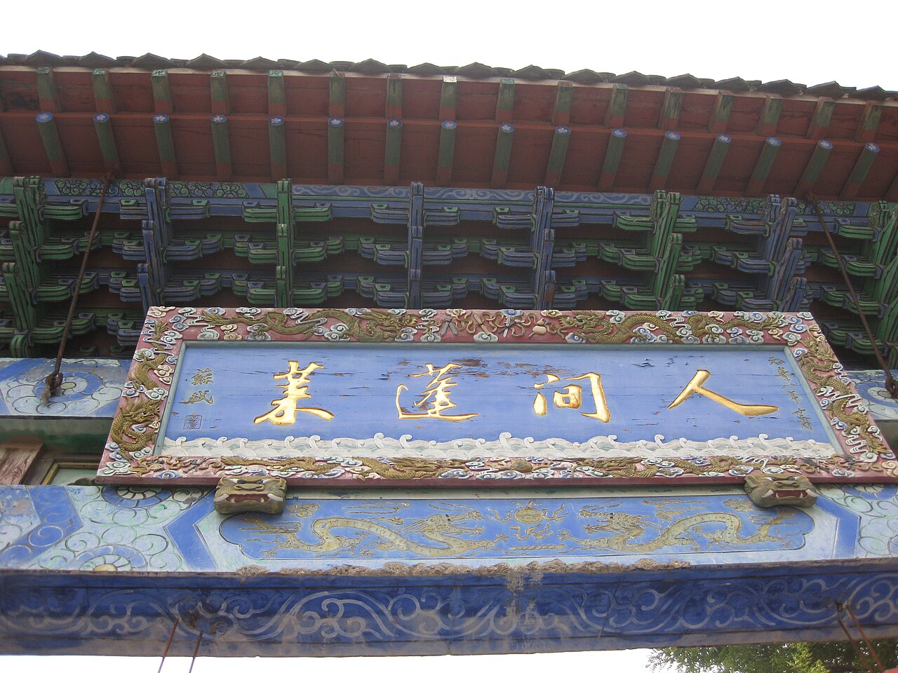 A blue and gold carved name board on a painted eave, under a tiled roof with green painted beams