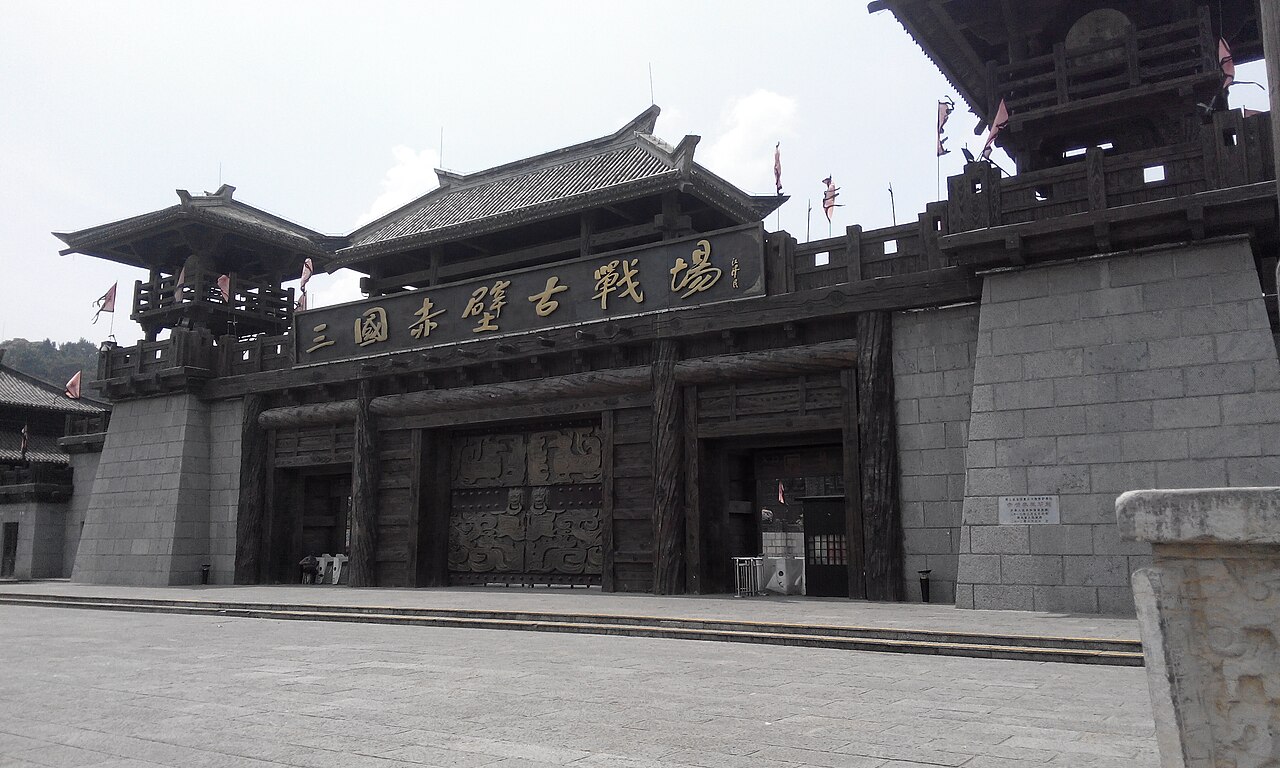 A broad grey stone gate building with dark timber upper storeys and a large horizontal plaque of gilded characters across its face, standing above a wide paved forecourt under a pale sky