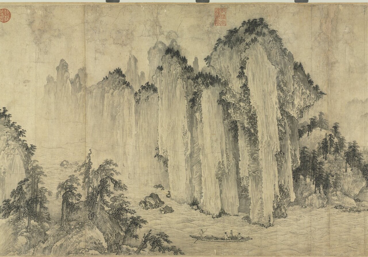 Detail of an ink handscroll painting showing tall stacked cliffs rising from a river bank, with pine trees and mist drawn in soft ink wash on aged paper