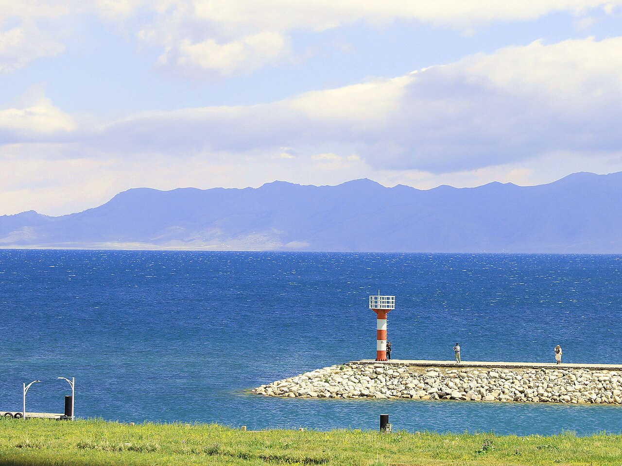 A small red and white lighthouse on a stone breakwater beside a very blue stretch of water, with a long mountain range behind under pale cloud