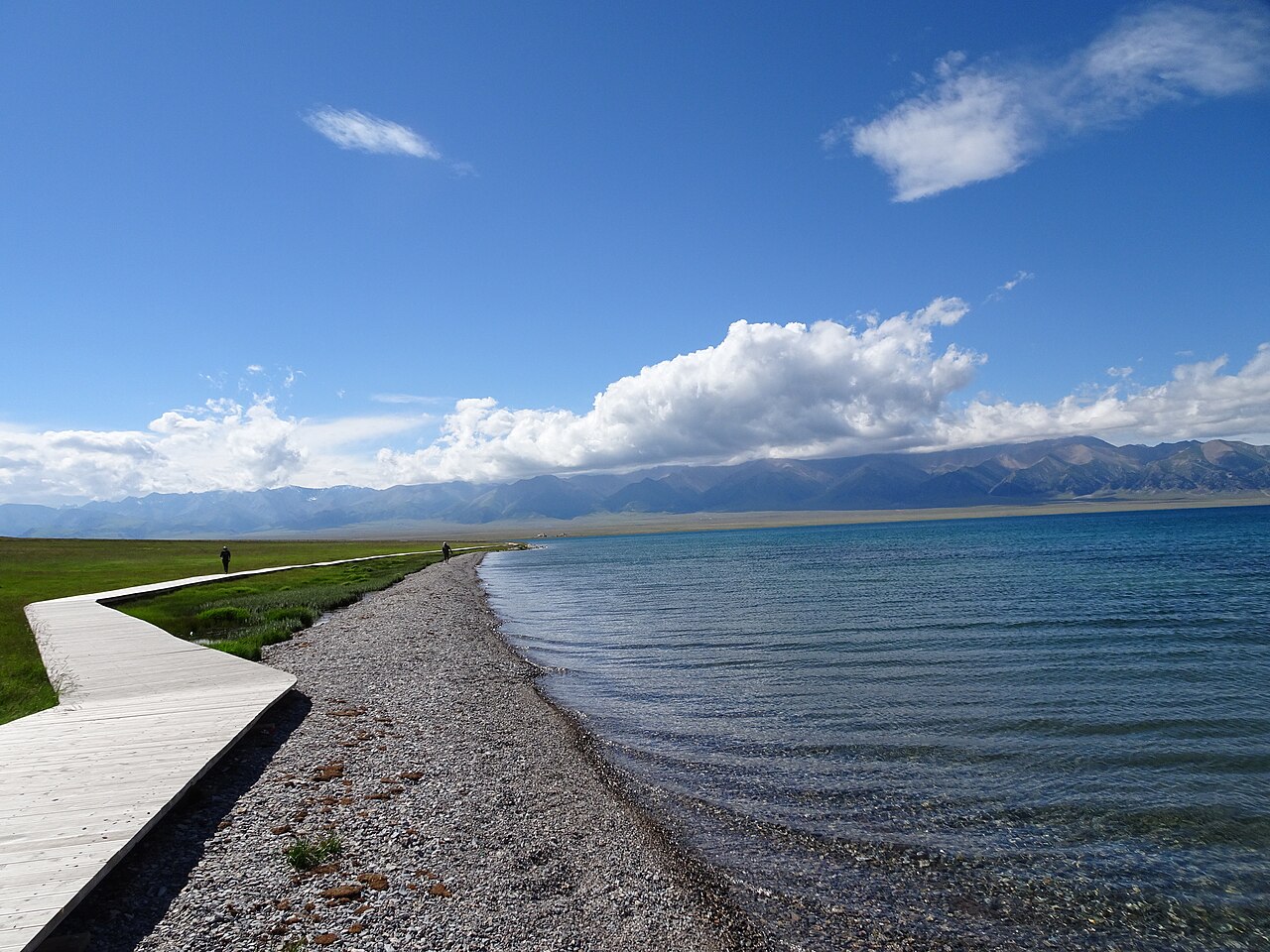 A gravel shore and a pale wooden boardwalk curving beside blue lake water, with grass at the left and a line of snowy mountains and white cloud on the horizon under a deep blue sky
