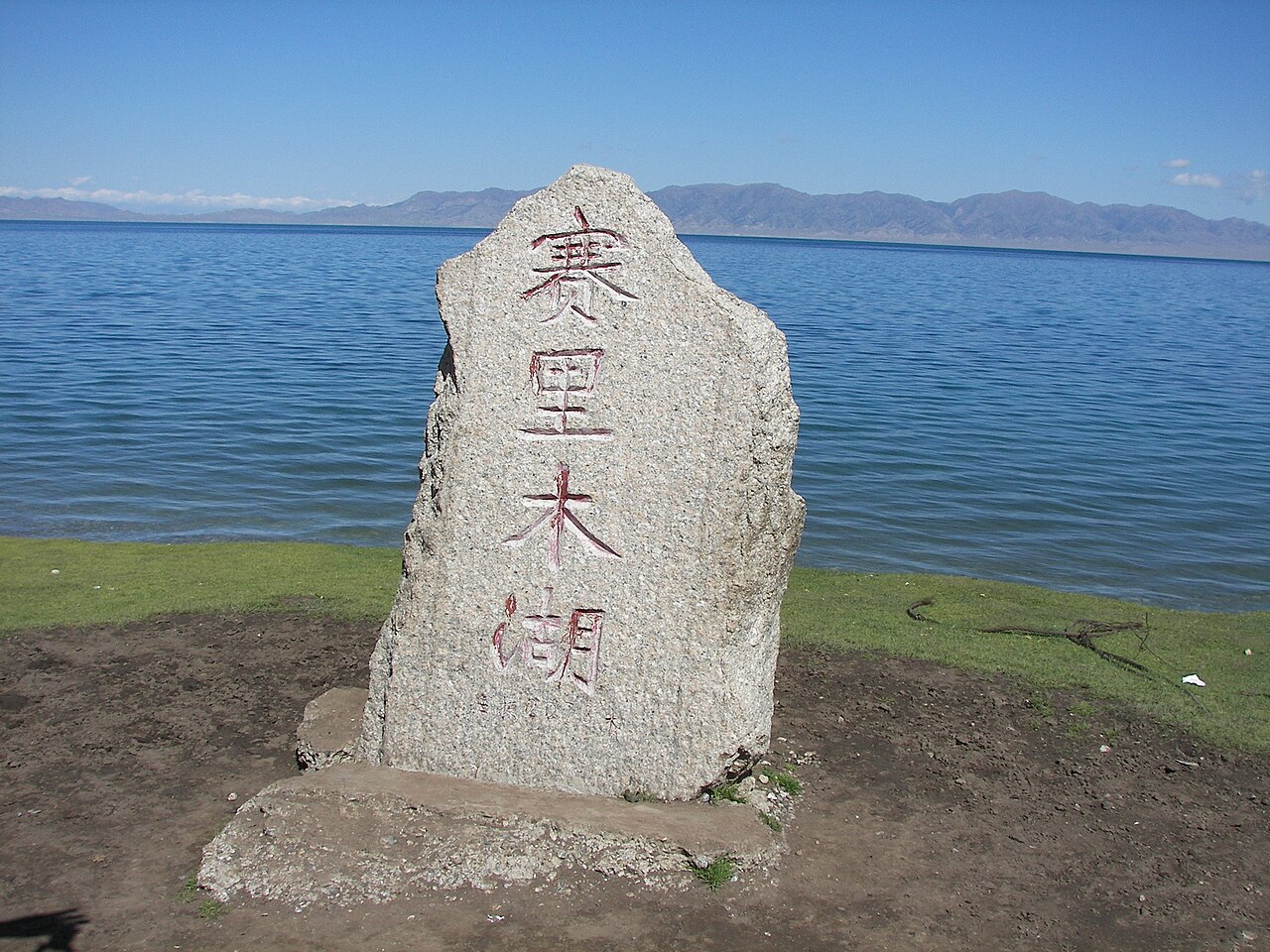 A grey stone marker carved with the red characters of the lake's name standing on a bare patch of ground beside the lake, with water and distant mountains behind