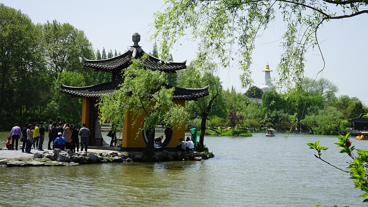 A small pavilion with two layers of eaves on a stone terrace at the lake edge, with willow branches hanging in front and visitors on the terrace under a pale blue sky