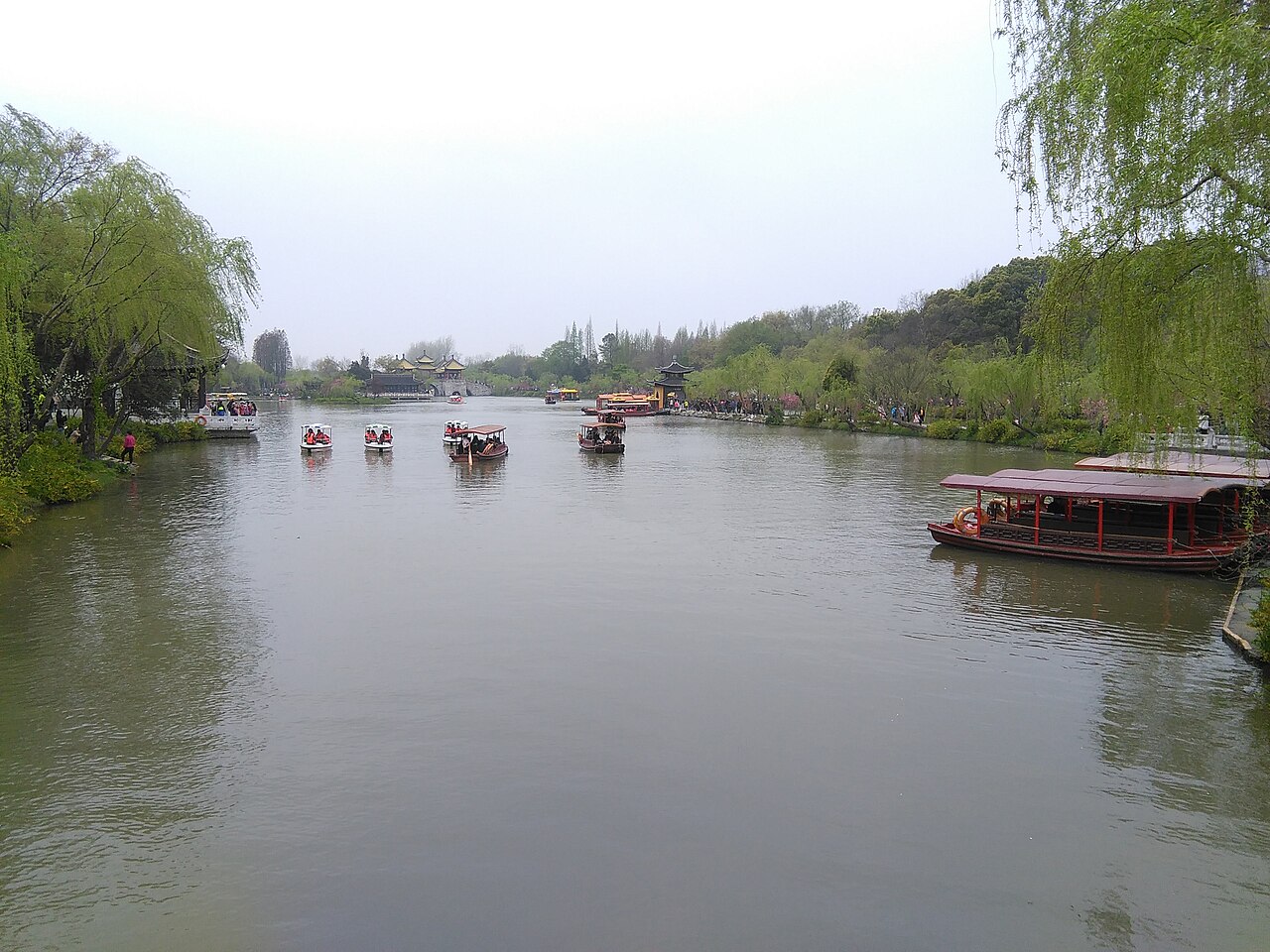 A broad calm stretch of lake with willows on the left bank, a pleasure boat moored on the right and several small boats moving in the distance under a pale sky