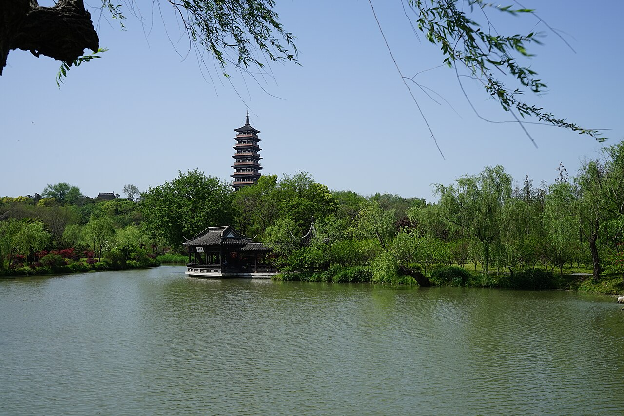 A white pagoda seen across still water over a bank of willows, with a small pavilion on a low island at the left and a blue sky above