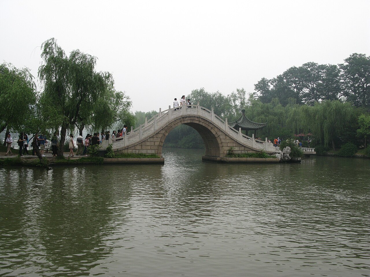 A pale stone arched bridge over a canal with willows on both banks under a grey sky