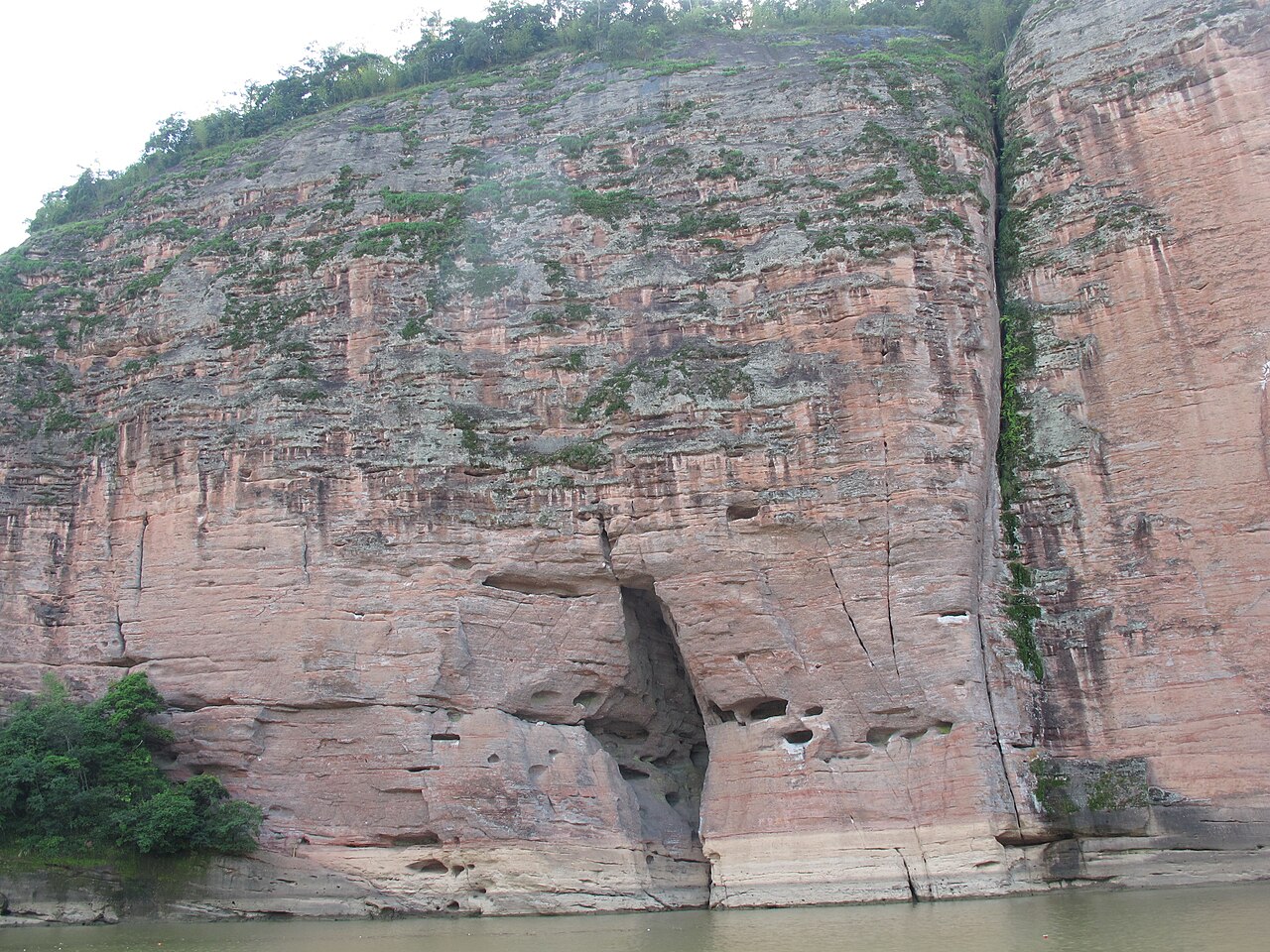 A close view of a wide red sandstone wall with a vertical fault line and a hollowed cave at its base, above calm water