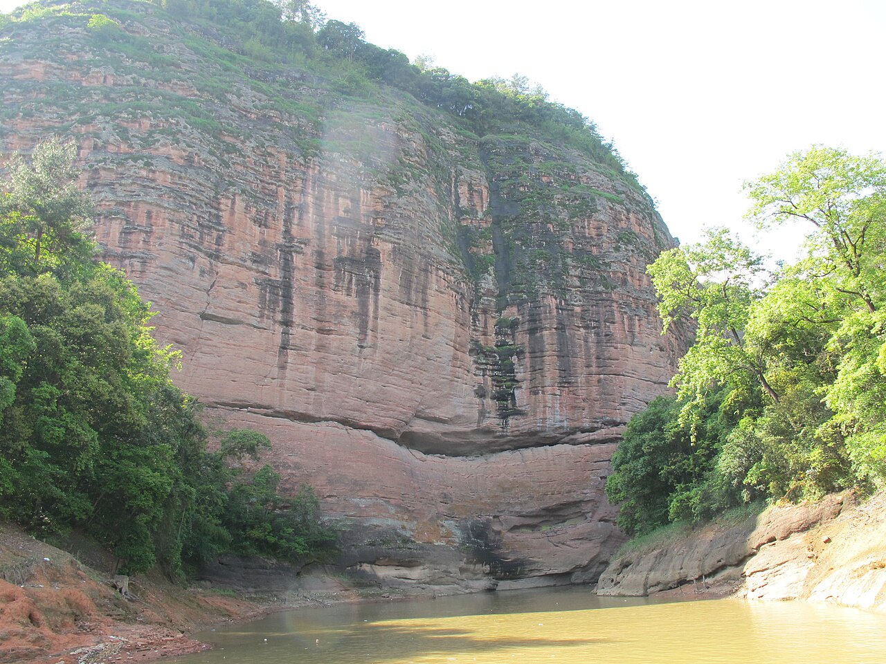 A huge pink-red sandstone cliff face streaked with green growth rising from the lake, with trees at the base and sky above