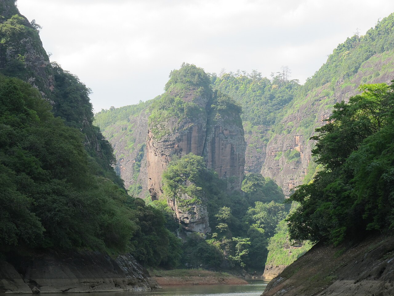 A narrow gorge between red cliffs and green forest, with a distant sandstone tower in the centre and bright sky above