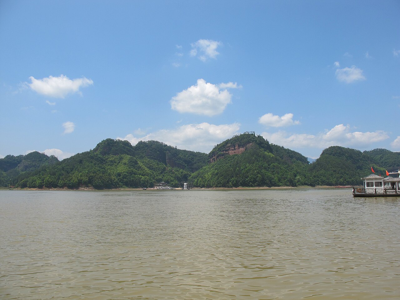 A wide lake with green rounded hills in the distance under a blue sky and a small tour boat at the right