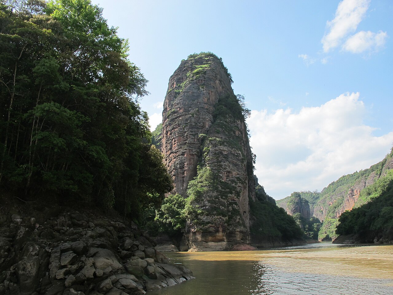 A tall red rock tower rising at a bend in a green creek, with dark trees on the left bank and white cloud in a blue sky behind