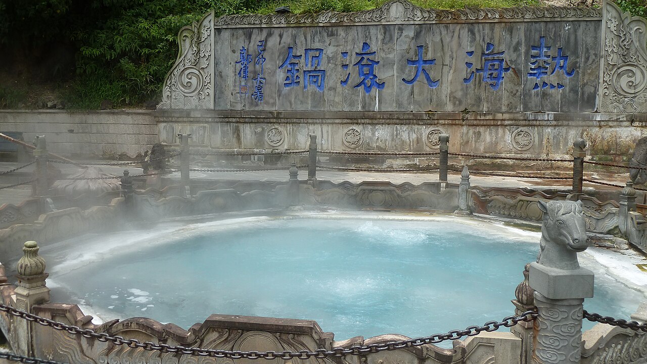 A round stone-edged hot spring pool with pale turquoise water giving off steam, with a carved stone dragon at its corner and a large carved name panel on the wall behind
