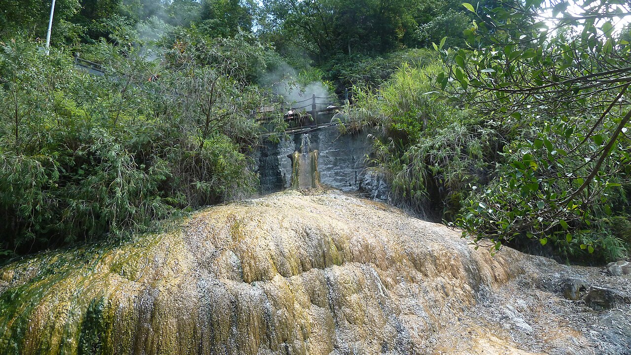 A broad mound of yellow and cream mineral deposit built up by spring water, with steam rising from a rock wall behind and green forest above