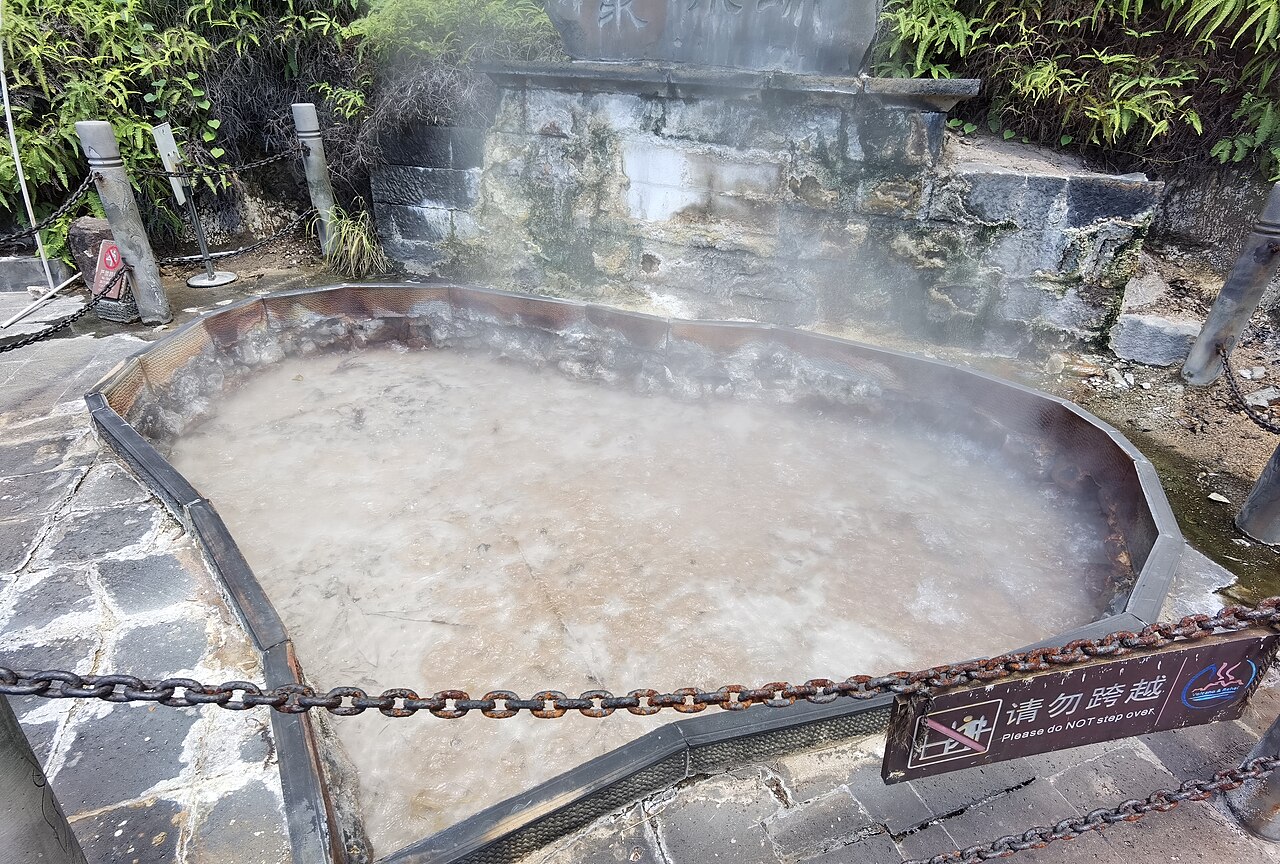A wide shallow spring pool ringed by a stone path and a low chain, with steam drifting across moss-covered rock