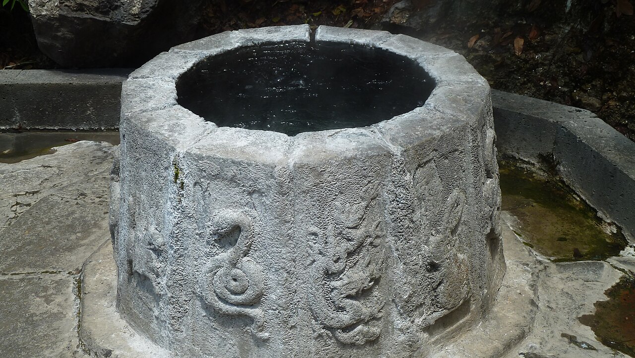 A carved stone well head with a dragon and a serpent in relief on its octagonal rim, beside a small channel of spring water