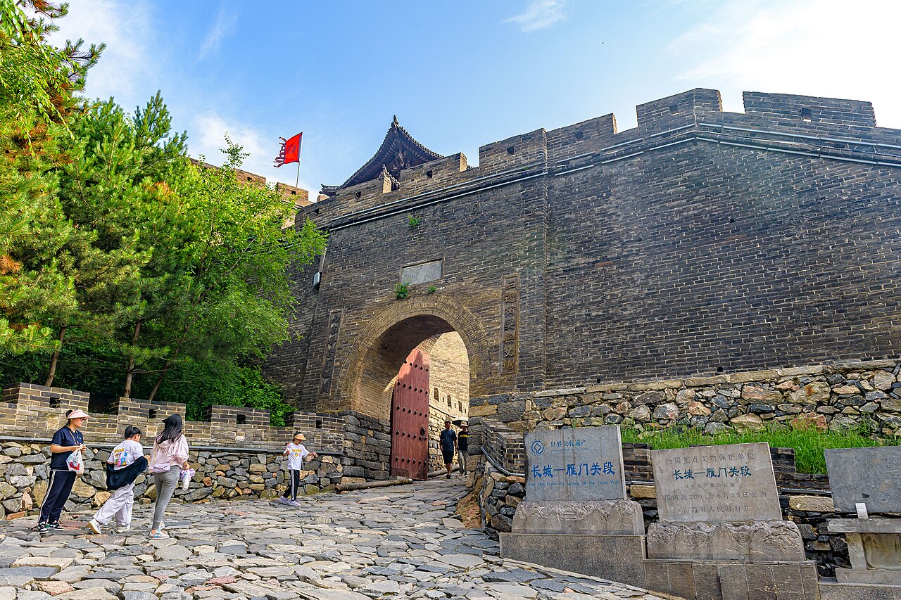 A brick barbican with an arched gate and red doors in a high wall, with visitors in front and a red flag above, with trees on the left