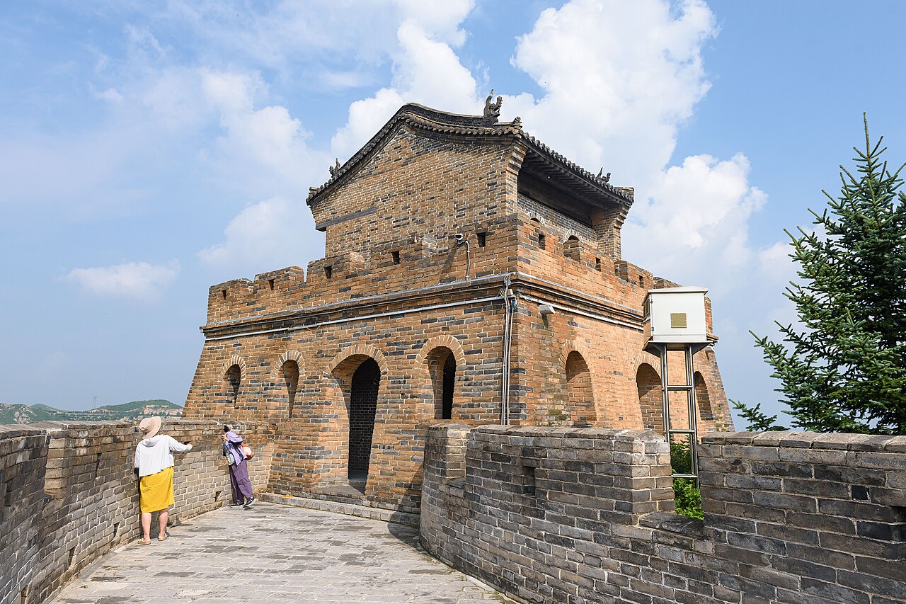 A square brick watchtower with arched openings on the wall walk, with two visitors on the paving and a wall running to the left under blue sky and white cloud