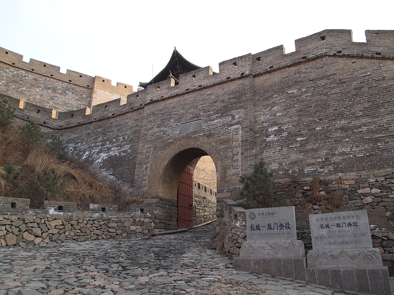 A grey brick gate with an arched opening and red door leaves, with two stone tablets standing in front on a cobbled path
