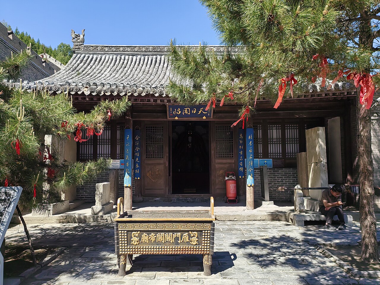 A small temple with a tiled roof and red painted pillars behind a stone incense burner, under old pines hung with red ribbons