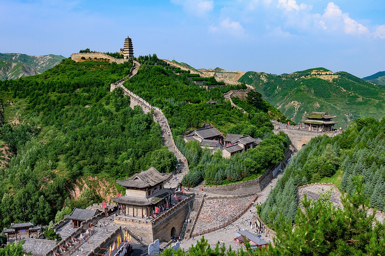 A stretch of the Great Wall climbing a green ridge from a brick-walled gate complex in the foreground, with a watchtower on the ridge top and bright hills beyond under a blue sky