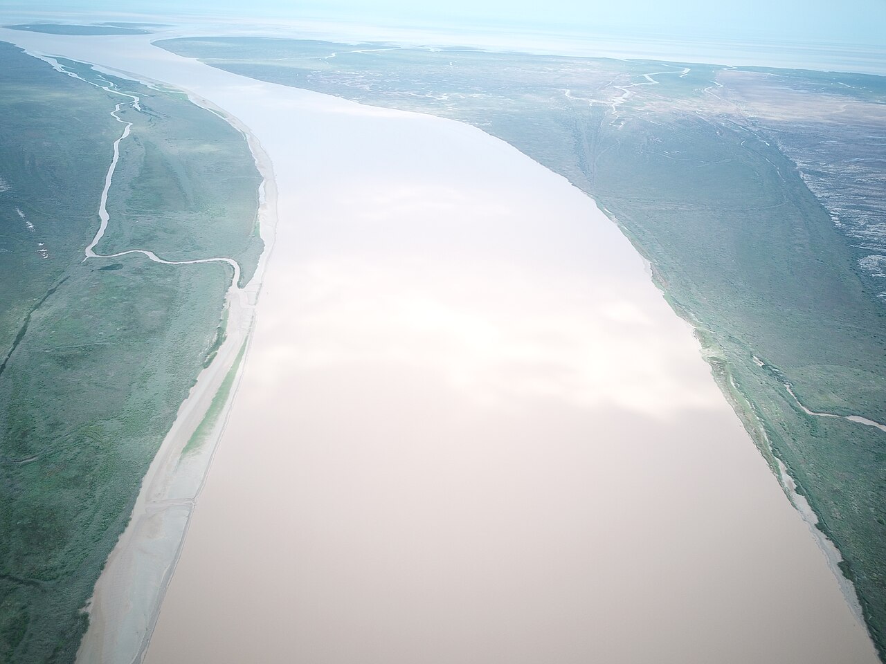 A wide pale brown river curving through green wetland banks seen from above, with small channels on the left bank