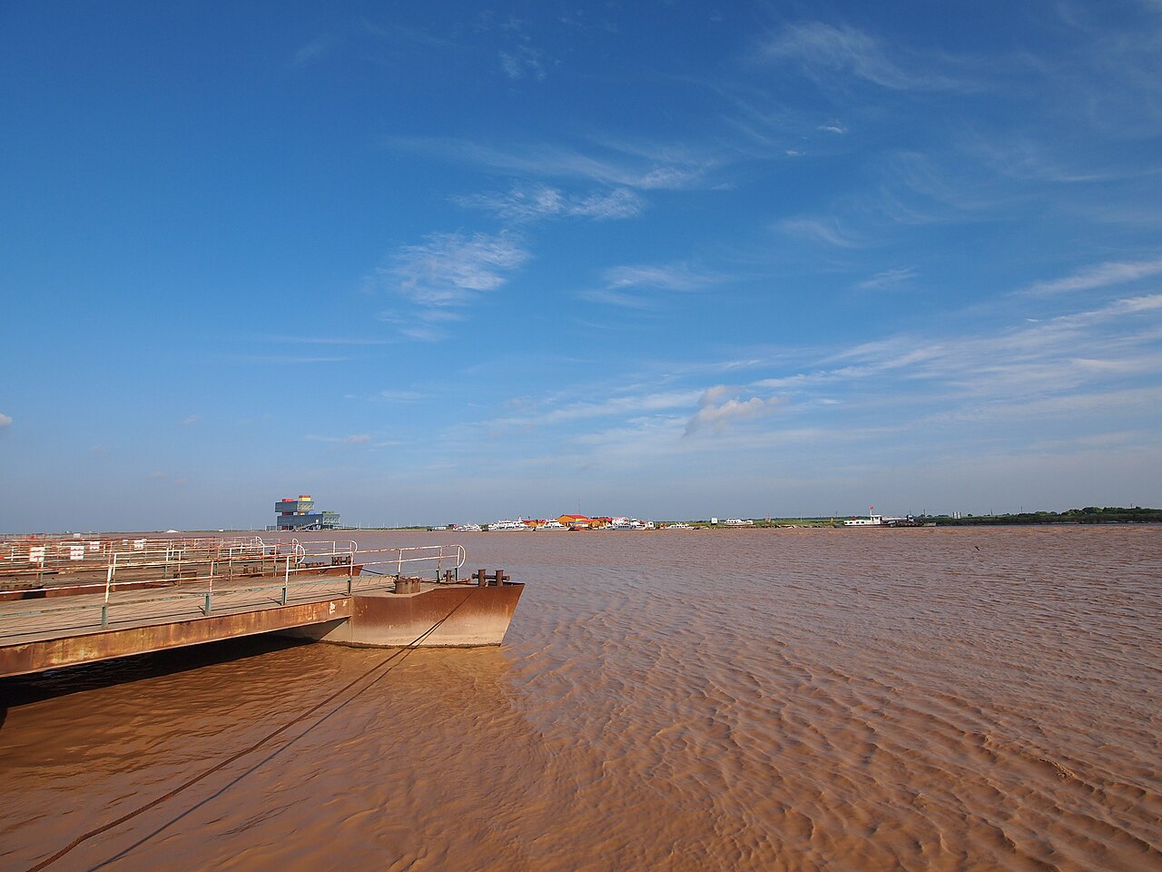 A wide brown river with a flat shore line, a moored barge and a long dock on the left, under a blue sky with thin white cloud