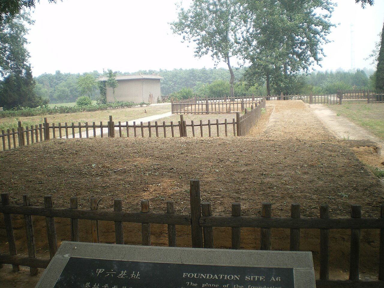 A raised earth platform behind a low timber fence, with a small round roof building and trees in the distance and a name plate in the foreground