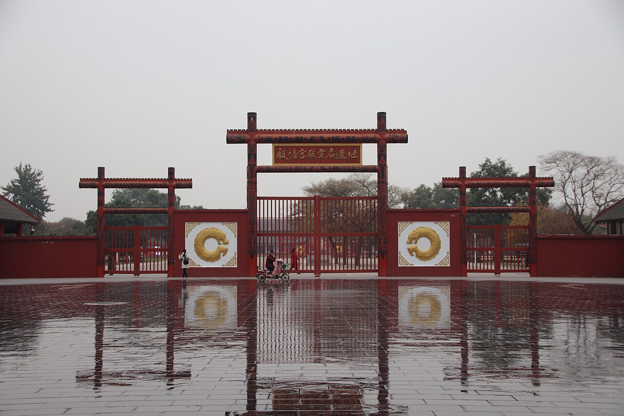 A red timber gate with three openings and gold circular ornaments set in a red wall, reflected in a wet paved court under a grey sky