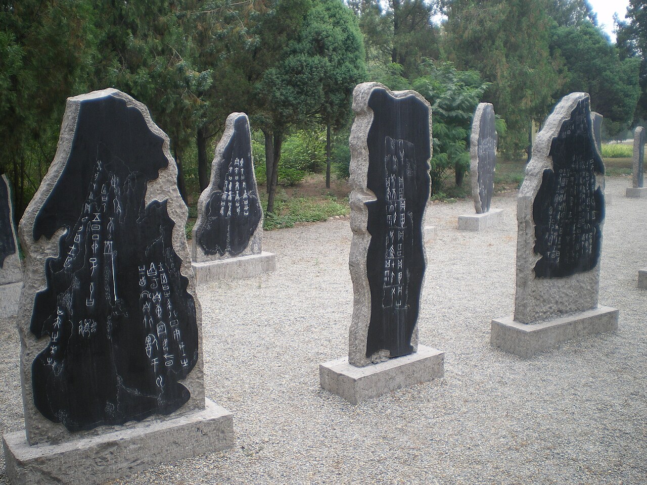 Several dark stone tablets shaped like irregular slabs carved with columns of characters, standing on low plinths on a pale gravel court under trees