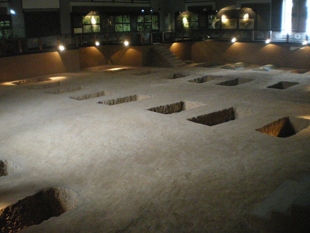 An excavated floor under a lit hall with rectangular pits and low earth dividers, seen from a gallery