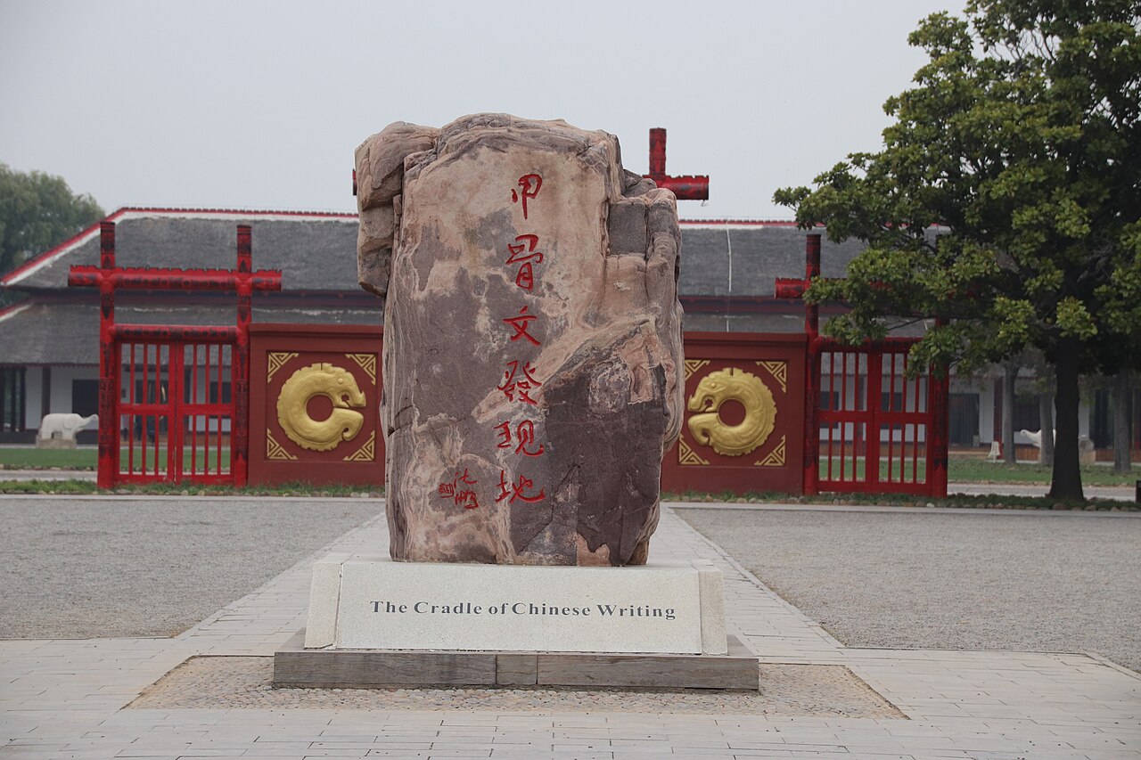 A weathered upright stone marker on a pale plinth carrying the English words The Cradle of Chinese Writing, with the red gate and low buildings behind it