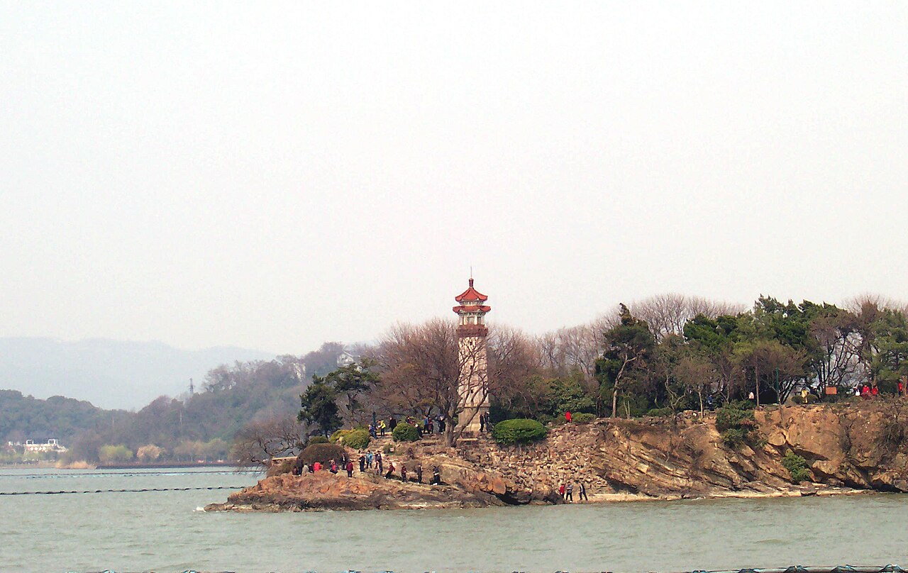 A small rocky islet with a red and white lighthouse and bare trees, in a calm lake with distant hills under a pale hazy sky