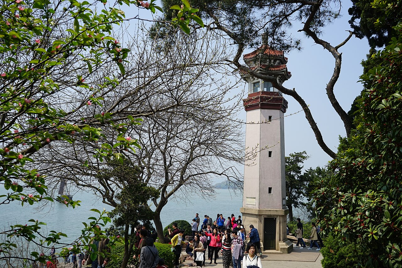 A white and red lighthouse tower seen from below through tree branches, with the lake and distant visitors behind