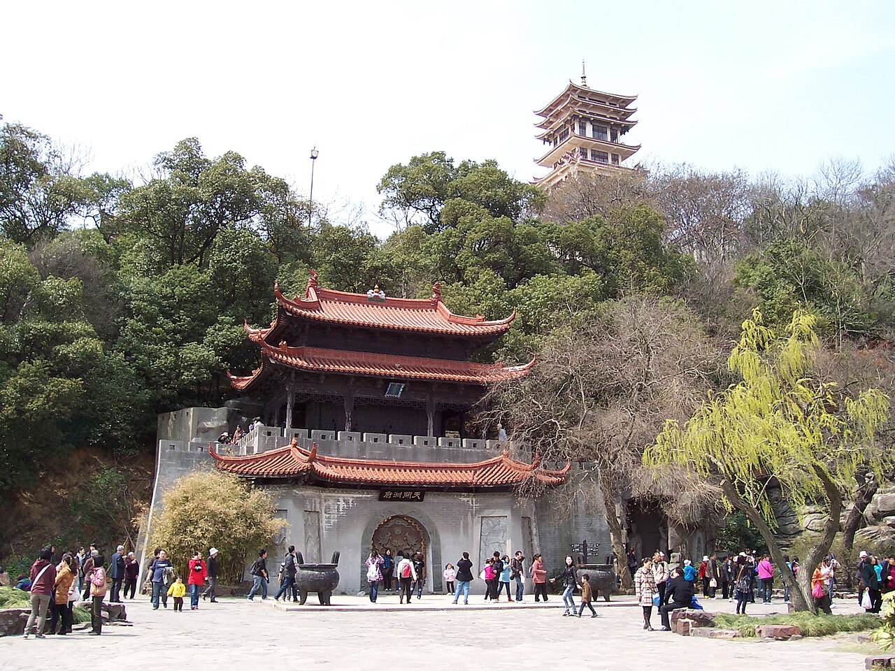 A temple pavilion with a red double roof among green trees and a tall tower on the hill behind it, with visitors on a paved court in front