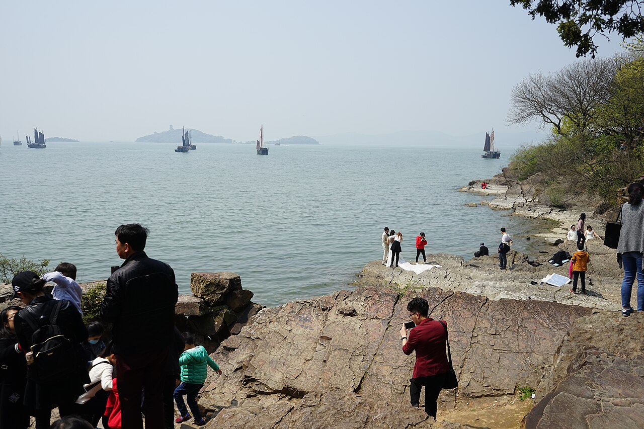 Visitors standing on a shelf of weathered rock at the water's edge, with islands and sailing boats on the lake behind