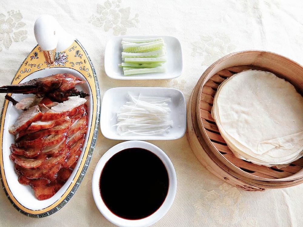 Sliced roast duck on an oval plate beside cucumber and scallion strips, a bamboo steamer of thin pancakes and a dish of dark sauce