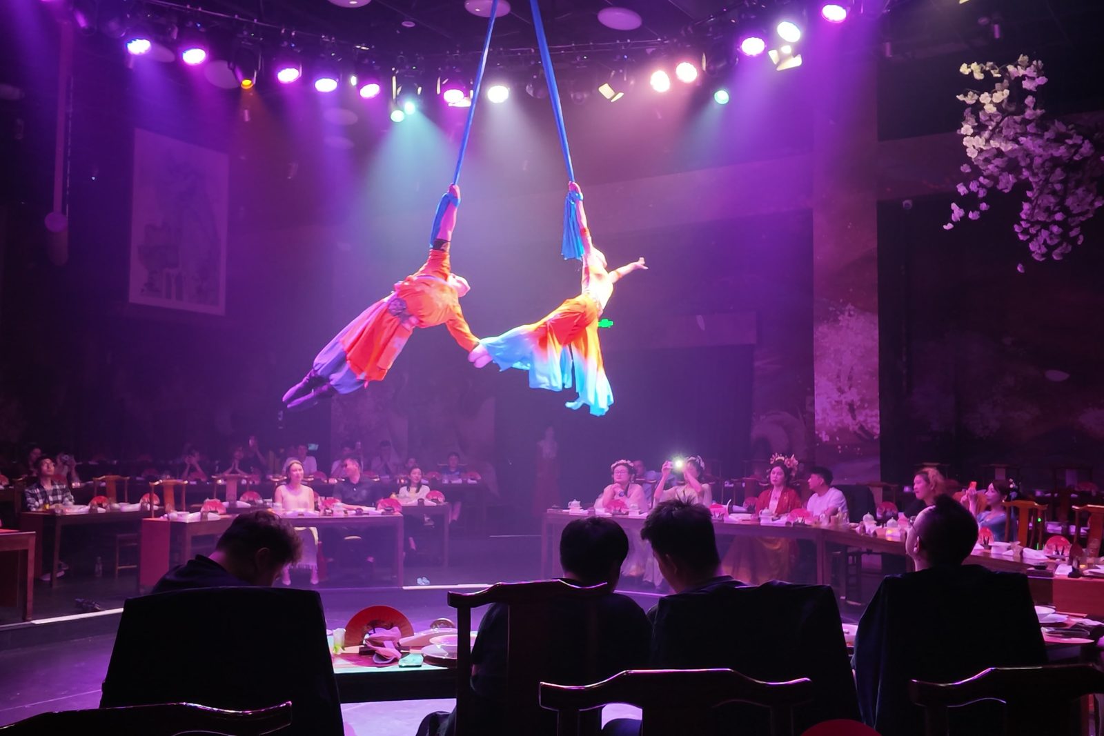 Two aerial silk performers in orange costumes above the tables of a banquet theatre in Hangzhou