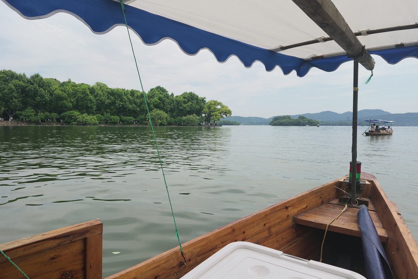 West Lake in Hangzhou seen from the bow of a wooden boat, with wooded islands and hills behind